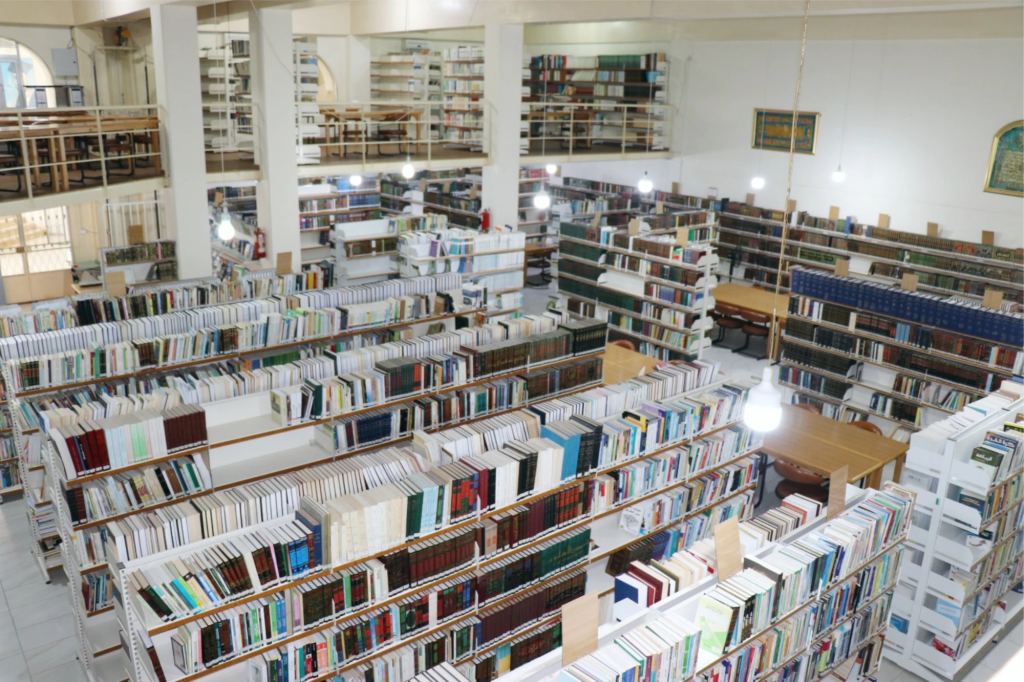 قاعة القراءة الرئيسية من الطابق العلوي: صفوف من الرفوف البيضاء تحمل كتبًا عربية ولاتينية، وبينها طاولات القراءة